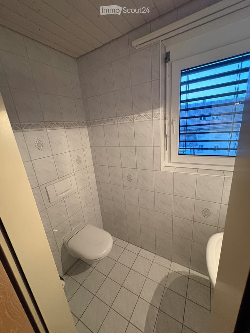 Small bathroom with tiled walls and floor, featuring a toilet and a sink, with a window providing natural light.