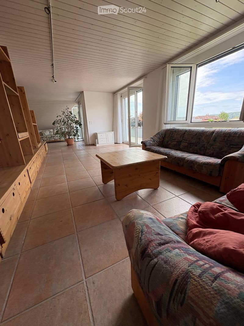 Bright living room with large windows offering a scenic view, featuring a patterned sofa, wooden coffee table, wooden cabinet, and a potted plant near the window.