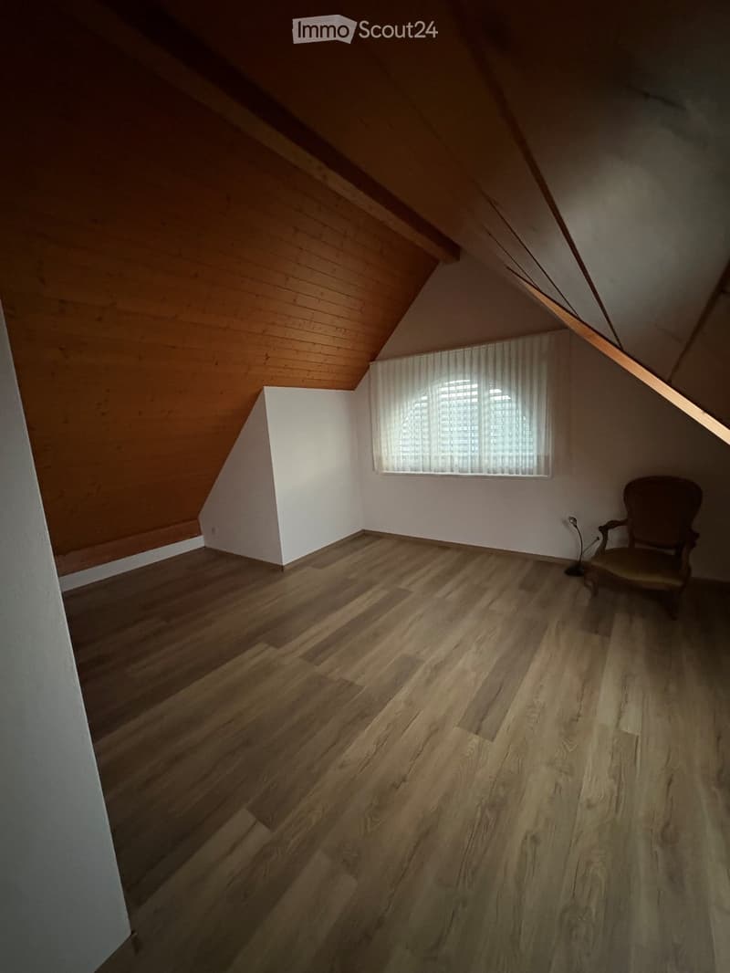An attic room with wooden floors, slanted wooden ceiling, white walls, and a single window with white blinds. A single wooden chair is placed near the window.