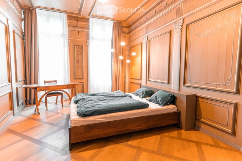 Elegant bedroom with wooden bed, matching nightstand, desk, chair, and large windows with curtains. Ornate wall panels and framed artwork.