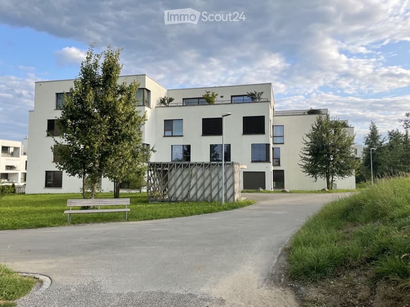 Modern white multi-story building with large windows and balconies, surrounded by greenery and a paved pathway.