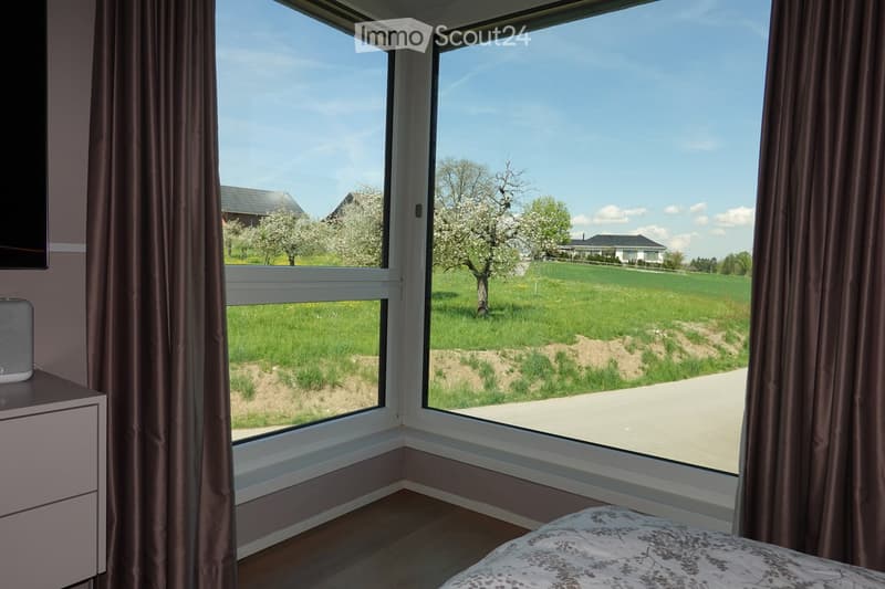 Bright bedroom with large windows offering a scenic view of a green field and trees. Features a bed with a patterned duvet, a nightstand with a speaker, and brown curtains.
