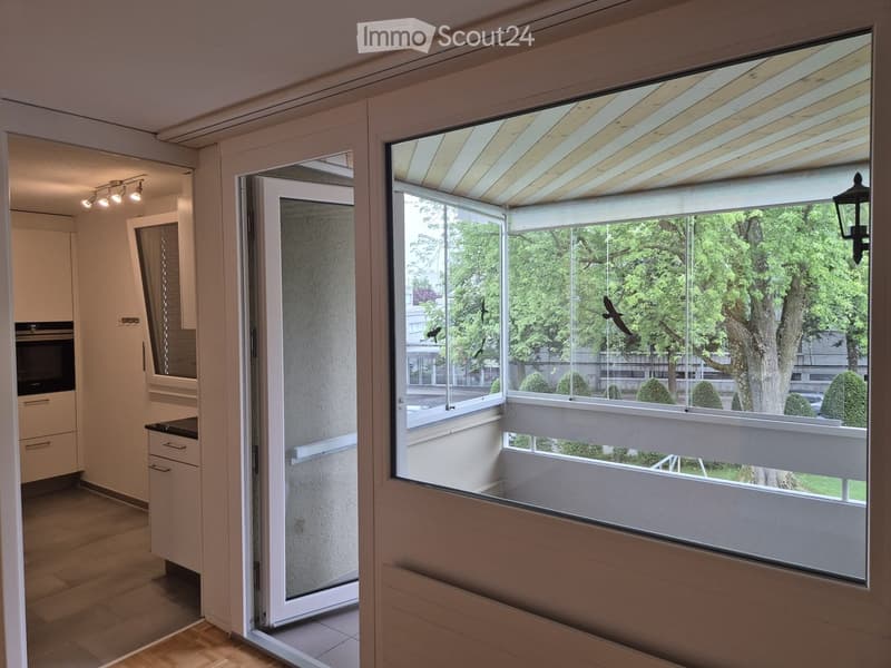 Balcony with large glass doors, wooden decking, and a view of greenery outside.