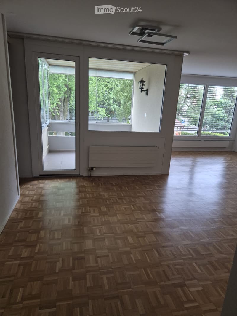 Empty room with wooden herringbone flooring, large windows, and a sliding glass door leading to a balcony.