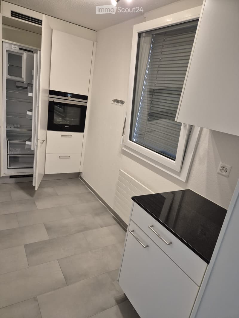Modern kitchen with white cabinets, black countertop, and stainless steel appliances including a refrigerator and oven. Features a window with blinds and tiled flooring.