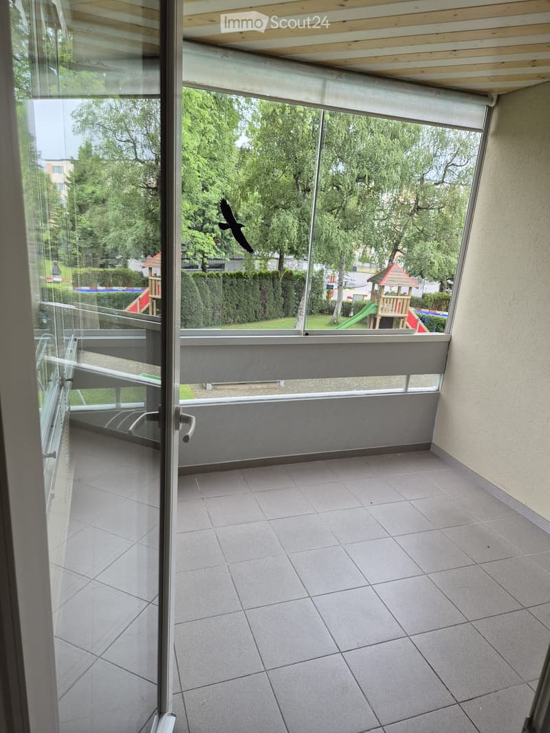 Balcony with tiled floor, large glass windows, and a view of a playground and trees.
