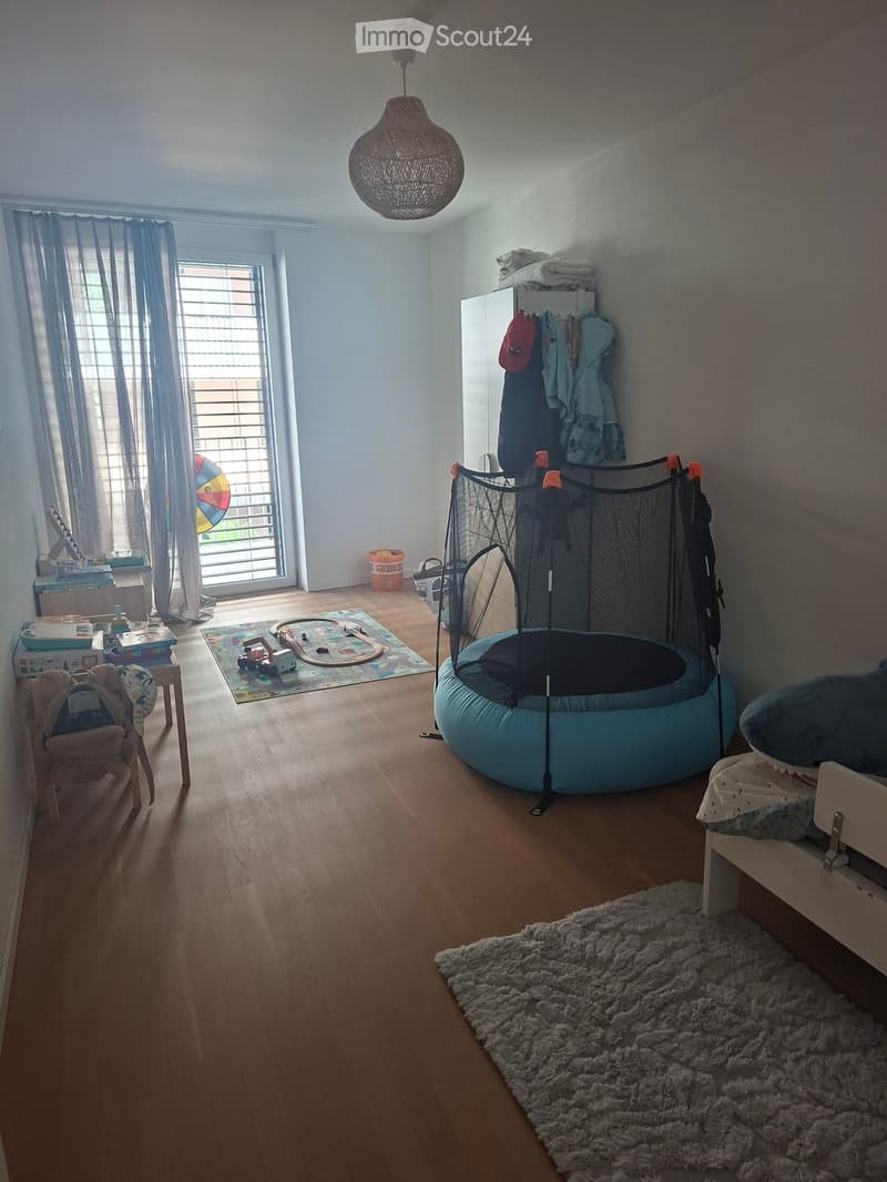 Child's room with wooden flooring, a padded bed, a toy train set on a mat, a small chair with a backpack, a wardrobe with clothes, a wicker hanging lamp, and a colorful balloon near the window.