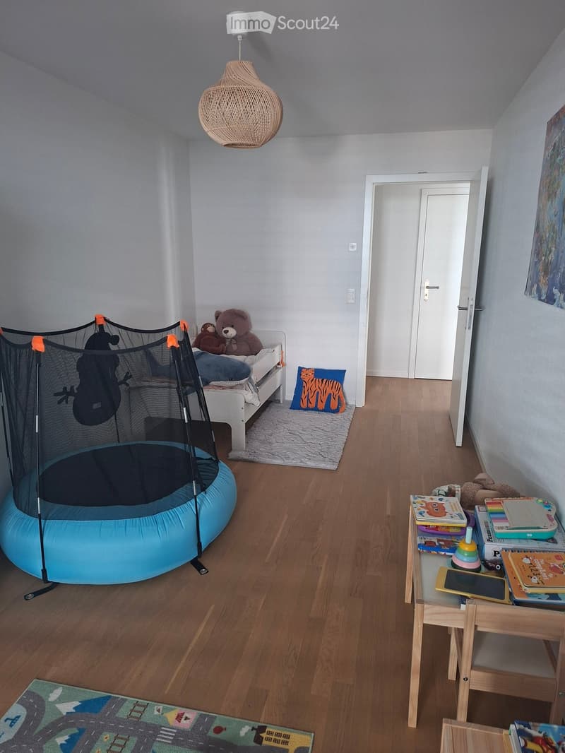 Child's bedroom with wooden flooring, a bed with stuffed animals, a small table with books and toys, a toy trampoline, and a colorful rug.