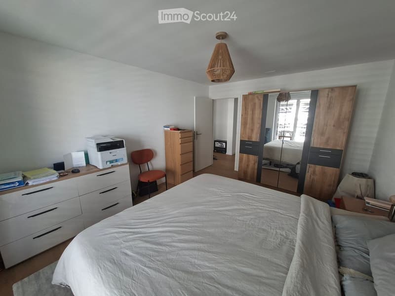 Well-lit bedroom with a large bed, a wardrobe with mirrored doors, a wooden chair, a printer on a drawer unit, and a hanging pendant light.
