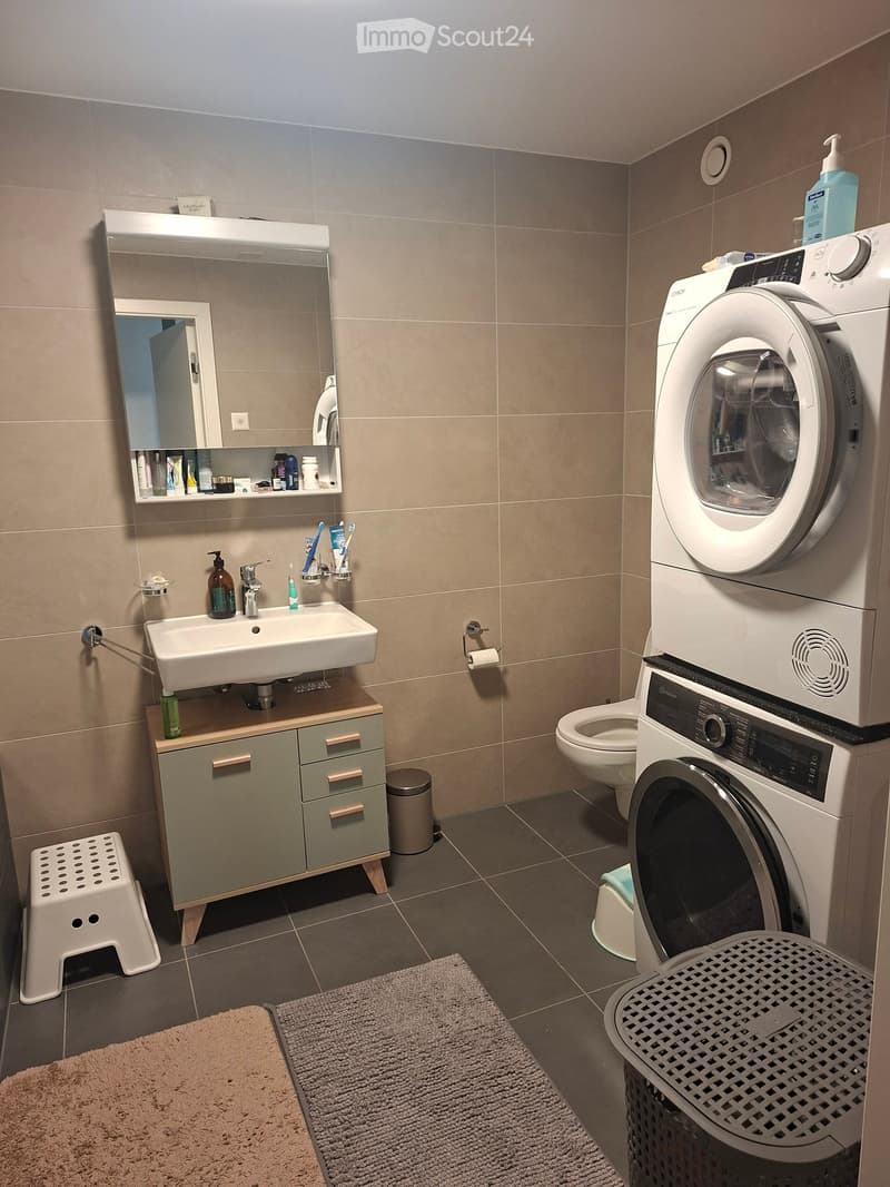 Modern bathroom with a sink, mirror, cabinet, toilet, and a stacked washer/dryer unit. Includes a small stool, toothbrush holders, and a soap dispenser.