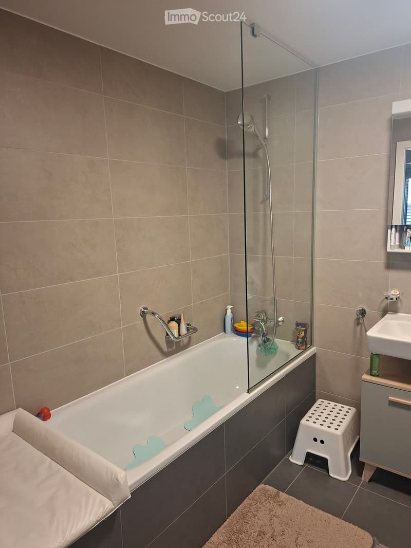 Modern bathroom with a bathtub, glass shower enclosure, sink, mirror, and a small white stool. Beige tiles and dark grey accents.