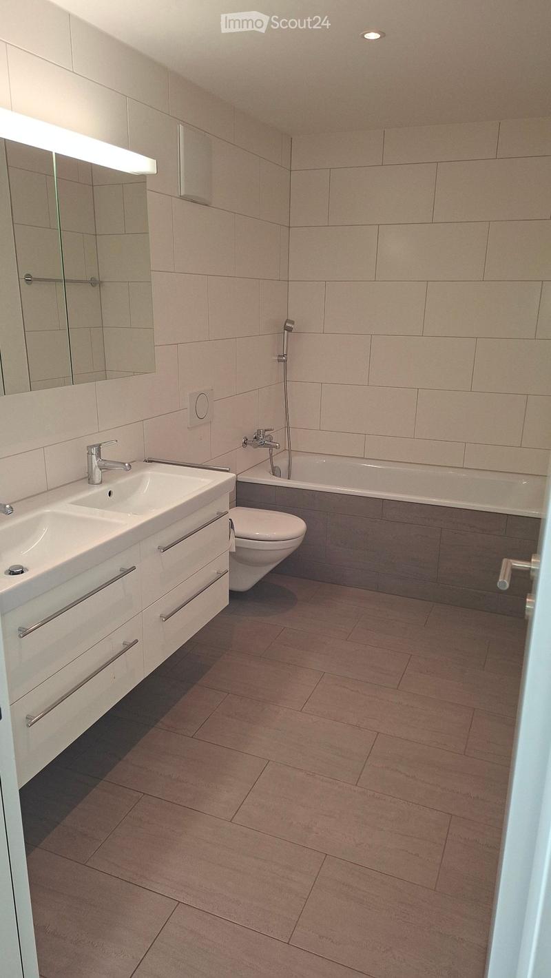 Modern bathroom with beige tiles, a double sink with chrome faucet, a toilet, a bathtub with a handheld shower, and a large mirror above the sink.