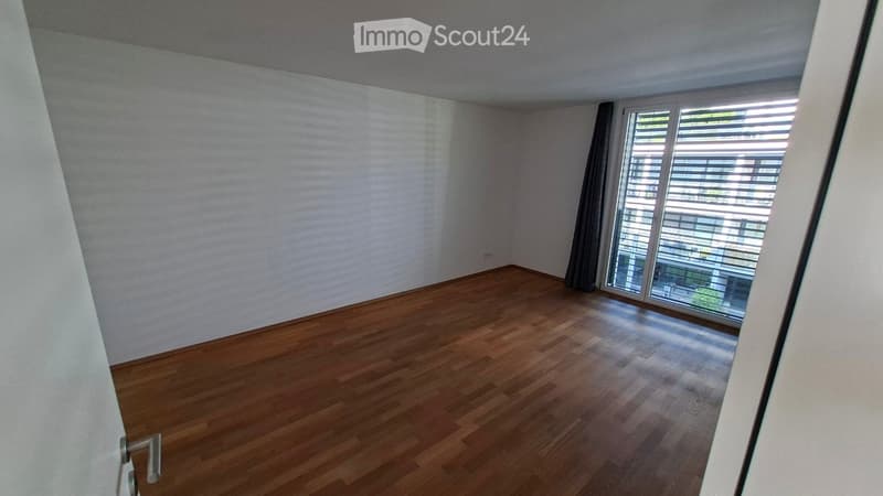 Empty room with wooden flooring, white walls, and large window with blinds leading to a balcony.