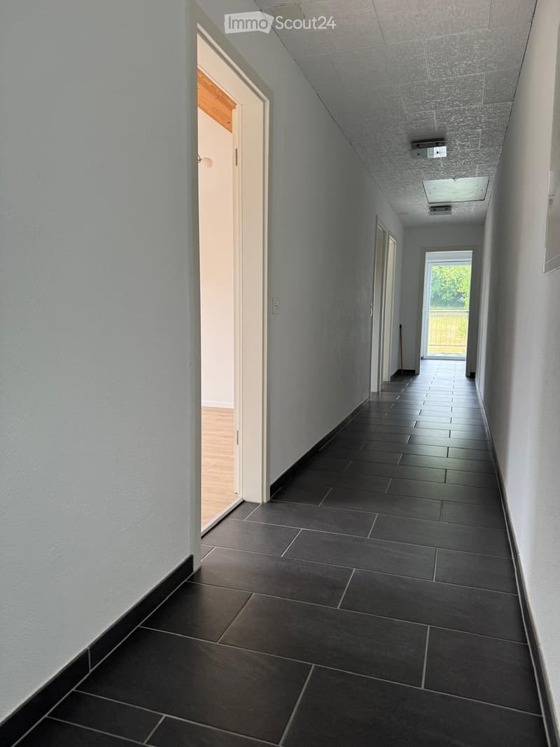 A long, narrow hallway with dark tiled flooring and white walls, featuring several doors and a window at the end.