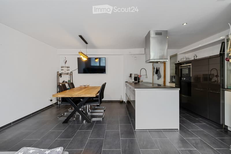 Modern kitchen with dark tiled flooring, wooden dining table, black chairs, flat screen TV, kitchen island with sink, modern faucets, cabinets
