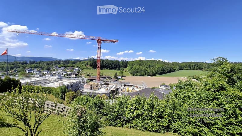 A construction site with a crane, partially built structures, and surrounding greenery and houses.