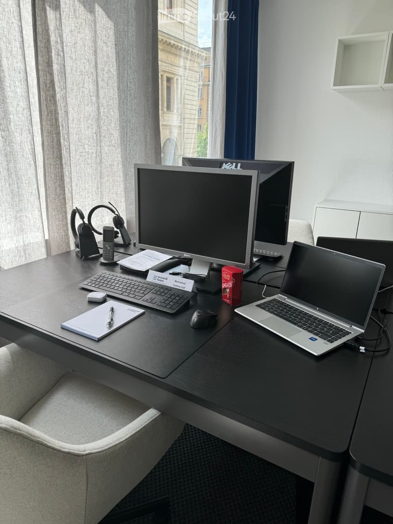 A modern, furnished room with a desk setup featuring a laptop, monitor, keyboard, mouse, and headphones. A notepad and pen are placed on the desk, and a comfortable chair is positioned in front of the desk.