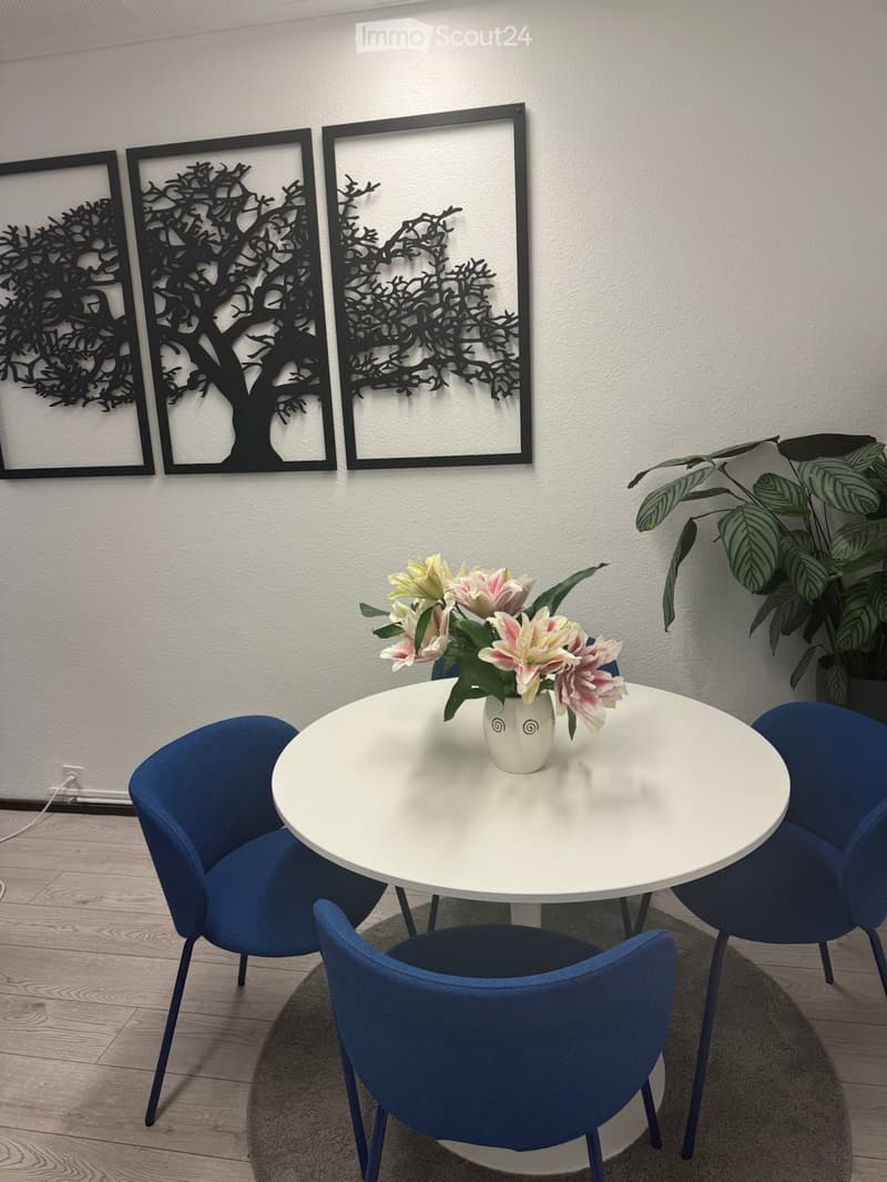 A modern dining area with a white round table, four blue chairs, a vase with pink and white flowers, and a leafy plant in the corner.
