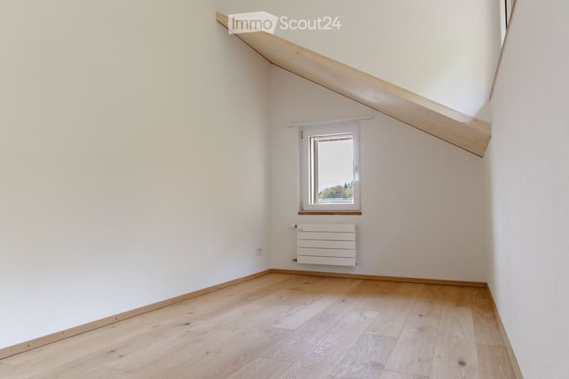 Empty attic room with wooden flooring, white walls, a single window, and a radiator. The sloped ceiling features exposed wooden beams.