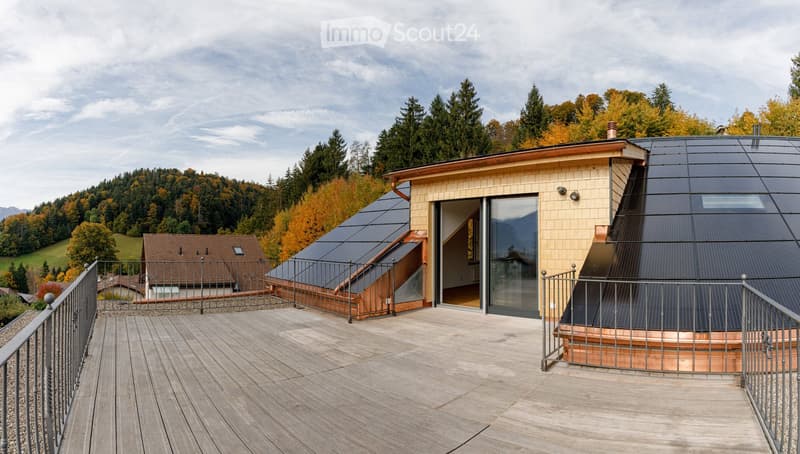 Modern house with a flat roof, solar panels, and large glass windows, surrounded by a wooden deck and autumnal forest.