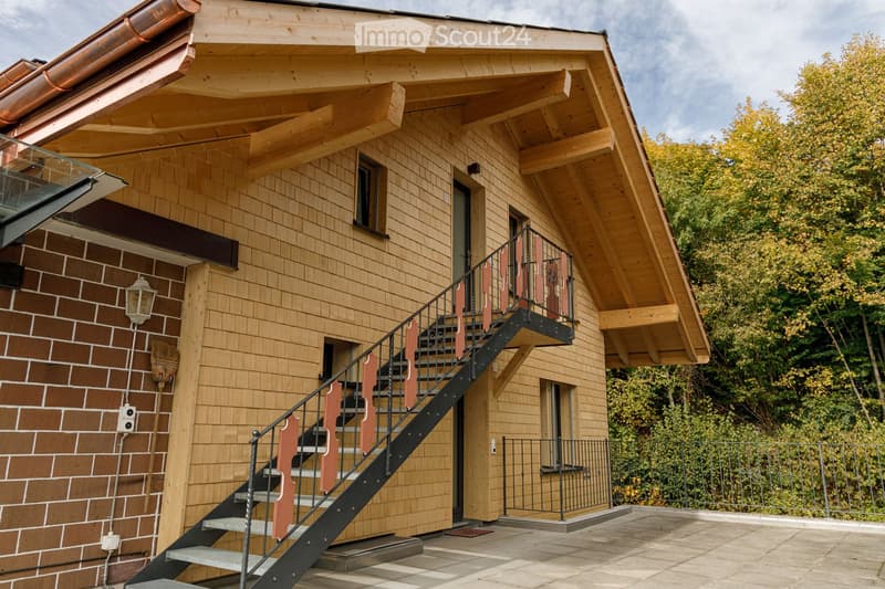 Modern two-story house with wooden exterior, brick section, raised ground floor, staircase leading to balcony, and surrounding greenery.