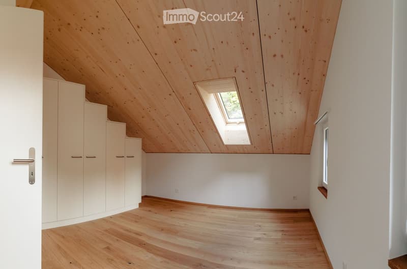 Empty attic room with wooden floor, white walls, built-in white cabinets, skylight window, and a small side window.