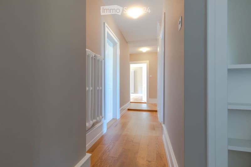 A long, narrow hallway with wooden flooring, beige walls, and white baseboards. The hallway features a radiator on the left wall and a thermostat on the right wall. The ceiling has recessed lighting, and the hallway leads to an open doorway at the end.