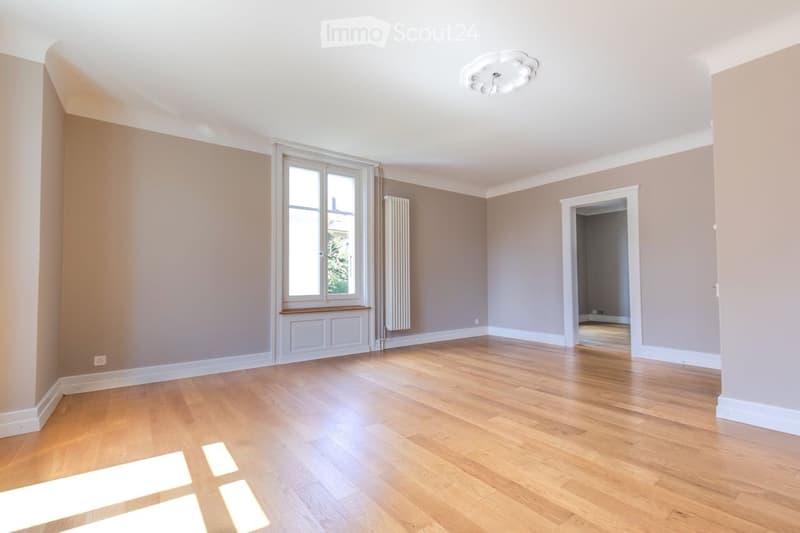 Empty room with wooden flooring, light-colored walls, large window, radiator, and a doorway leading to another space.