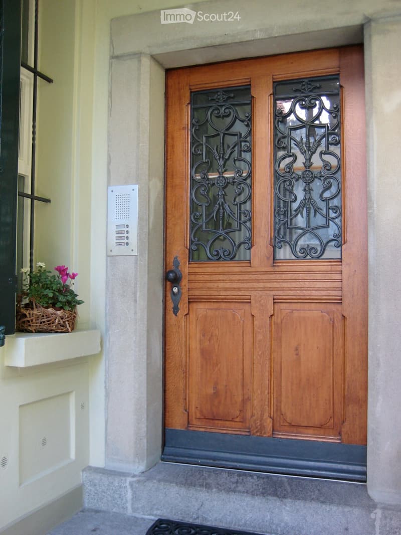 Wooden front door with decorative glass panels, intercom system, and a small flower basket on the left side.