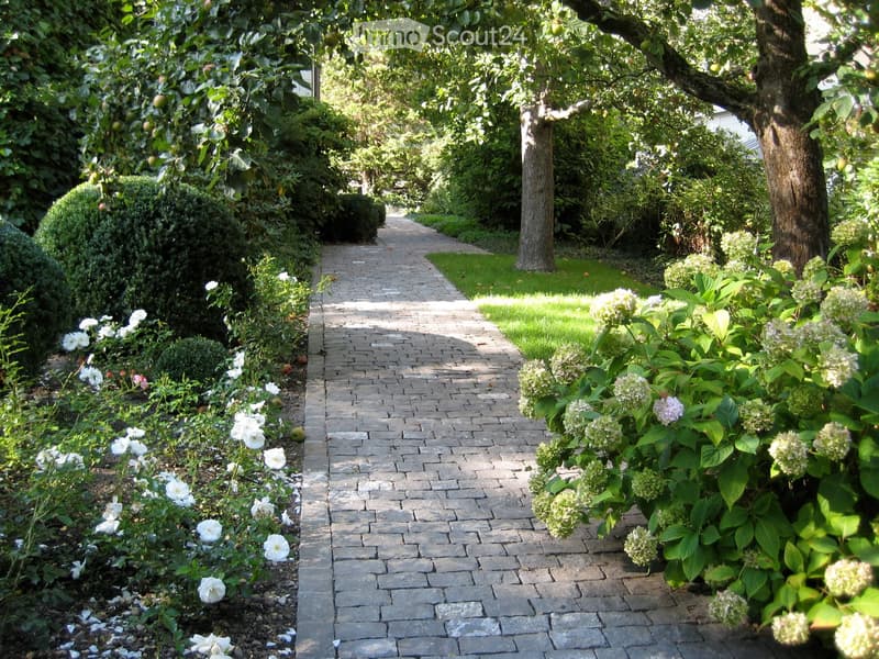 A well-maintained garden pathway lined with neatly trimmed hedges, flowering shrubs, and trees, creating a serene and inviting outdoor space.