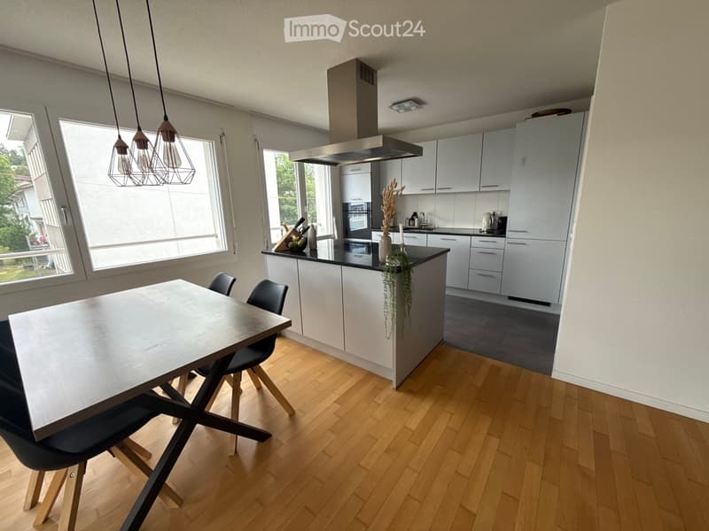 Modern kitchen with white cabinets, a central island, and a dining area. Features include pendant lights, a stainless steel range hood, and a black countertop.