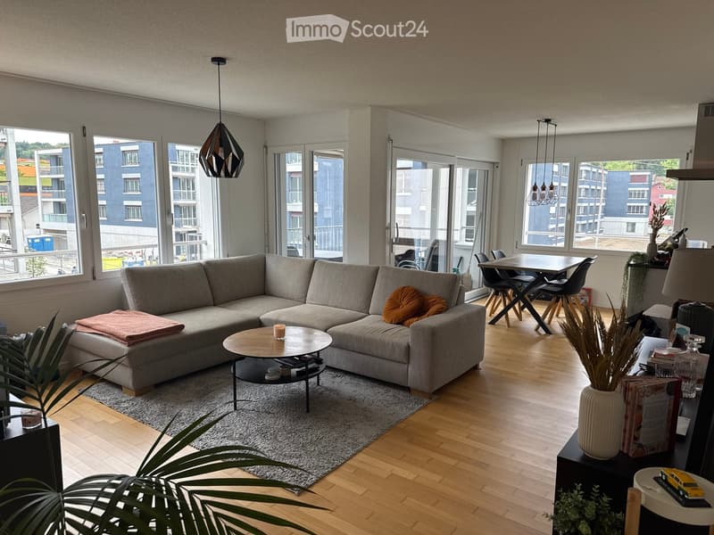 Modern living room with large windows, grey sectional sofa, coffee table, side table, dining area with chairs and table, decorative plants, and lamps.