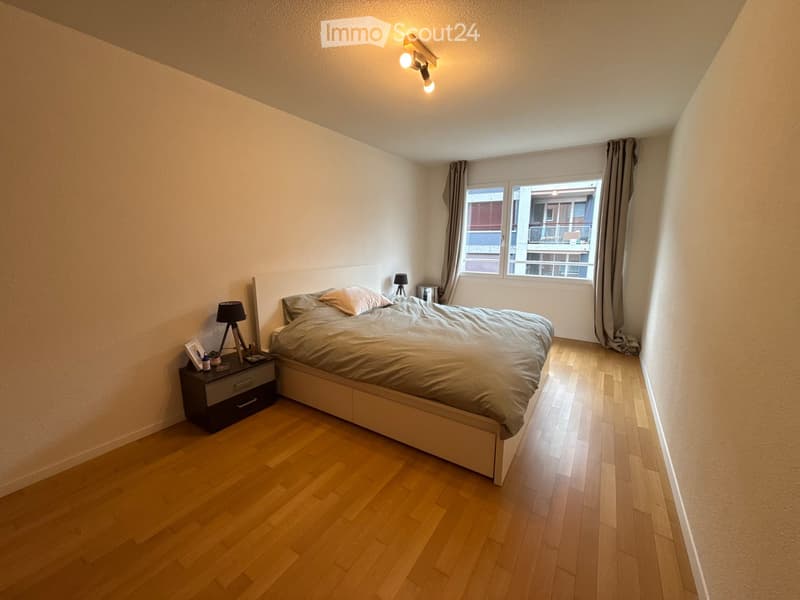 Modern bedroom with wooden flooring, a large bed with grey bedding, a nightstand with a lamp, and a window with curtains.