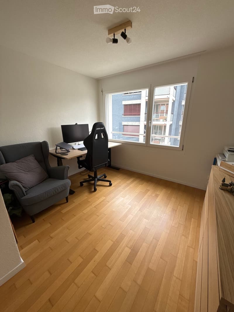 A modern, well-lit room with wooden flooring, a large window offering a city view, a comfortable sofa, a desk with a laptop, and a gaming chair.
