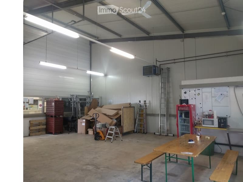 A spacious, industrial-style break room with a large table and benches, a Coca-Cola refrigerator, microwave, and various storage items including ladders and cardboard boxes.