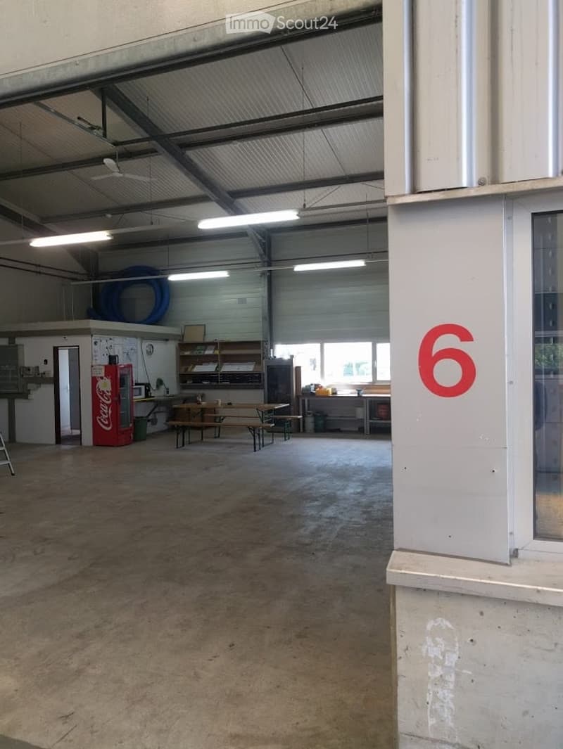 The image shows a spacious, open room with a concrete floor and high ceilings. The room features industrial-style lighting and exposed metal beams. There is a small kitchenette area with a refrigerator, a bench, and a table with chairs. The room also incl