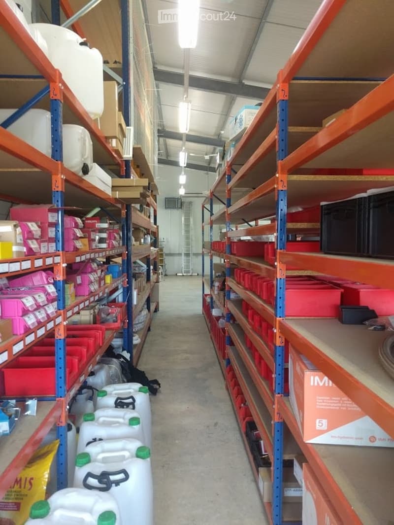A well-lit storage area with metal shelving units filled with various items including plastic containers, storage bins, and boxes. The floor is concrete, and the ceiling has exposed beams and fluorescent lighting.