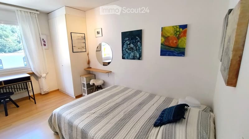 Cozy bedroom with a double bed, nightstand, mirror, and artwork on the walls. The room features wooden flooring and large windows with white curtains.