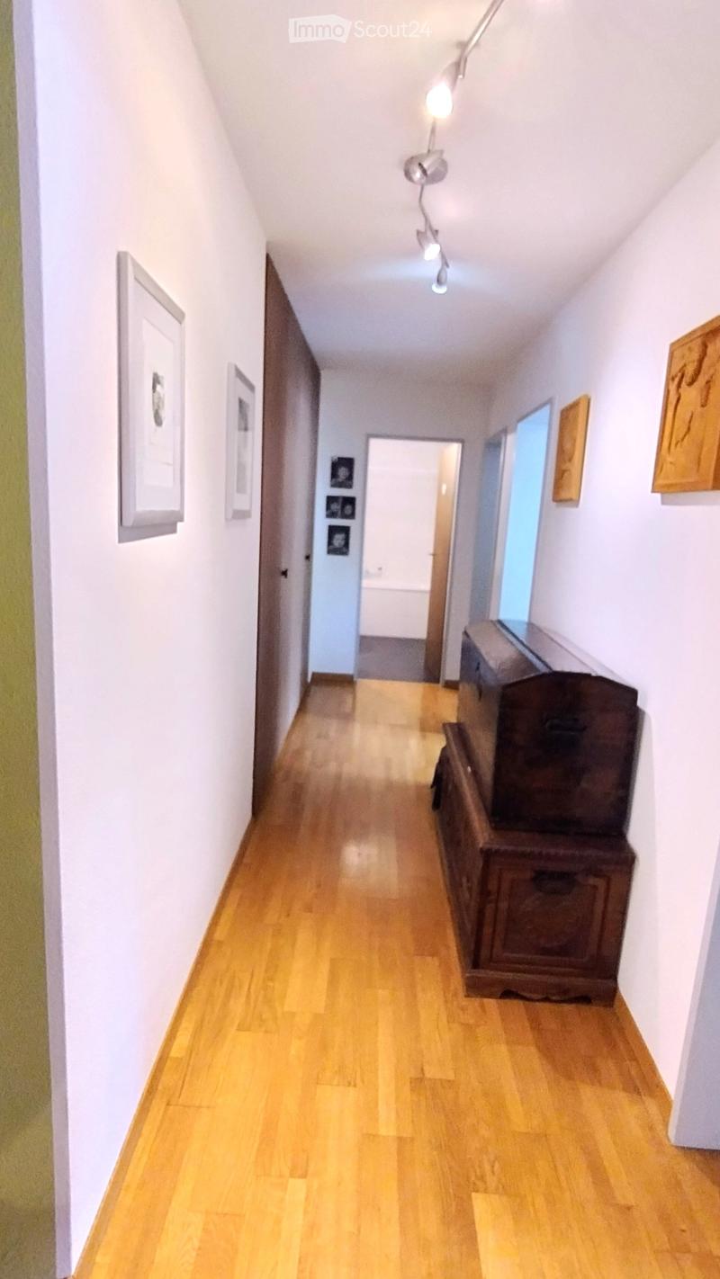 A narrow hallway with light wooden flooring, white walls, and several framed pictures. A small wooden cabinet is placed on the right side, and a doorway leads to another room at the end.