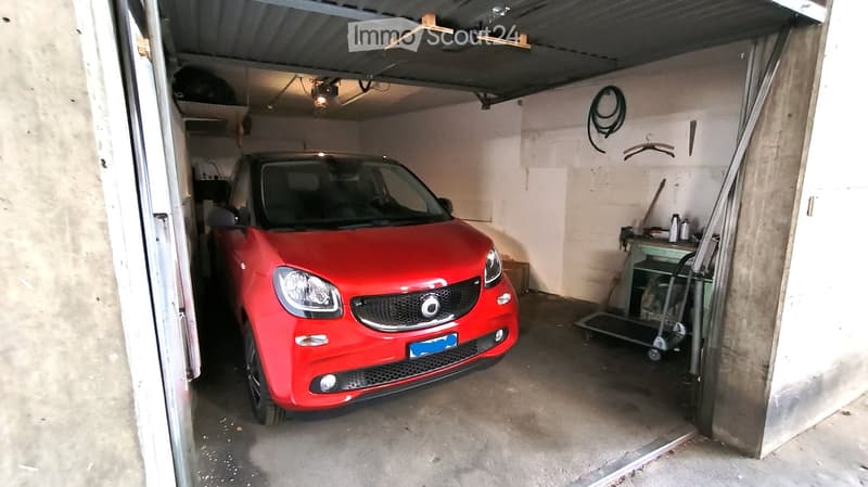 Garage with a red car parked inside, concrete floor, white walls, and storage items on the right side.