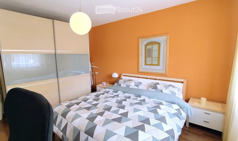 Modern bedroom with orange walls, a bed with geometric patterned bedding, a nightstand with a lamp, a wardrobe, and a framed picture on the wall.