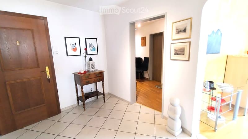 Hallway with tiled floor, wooden door, small wooden table with decorative items, framed pictures on walls, and a glimpse of a dining area with chairs and a glass table.