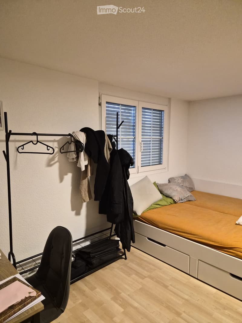 Small bedroom with a single bed, a chair, a clothing rack, and a table with books.