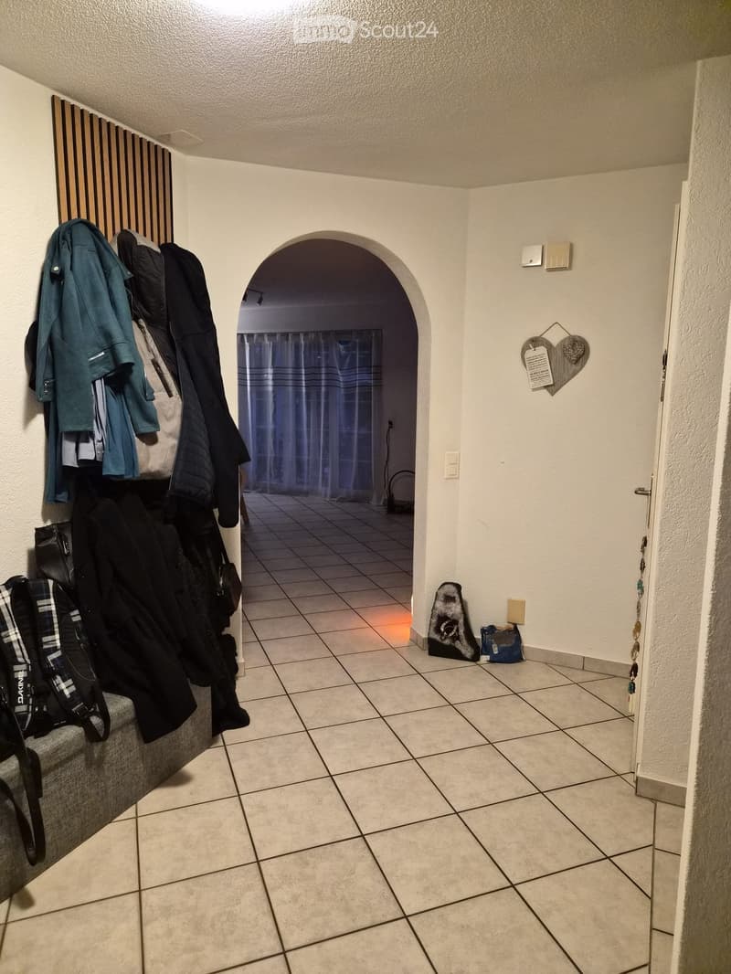 Hallway with tiled floor, arched doorway leading to another room, coats and bags hanging on the wall, small decorative item on the wall.