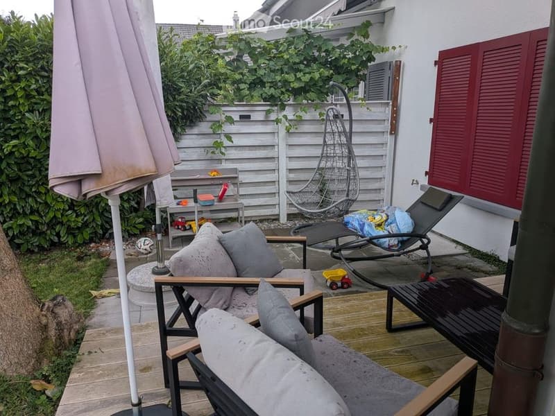 A cozy outdoor patio area with a wooden deck, lounge chairs, a small table, and a hanging egg chair. There is a large umbrella, a small table with toys, and a lush green hedge in the background.
