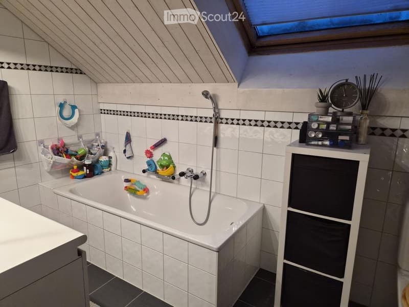 A cozy bathroom with a bathtub, showerhead, storage cabinet, clock, potted plant, and various toiletries and toys visible. The room features white tiled walls and a slanted ceiling with a skylight.