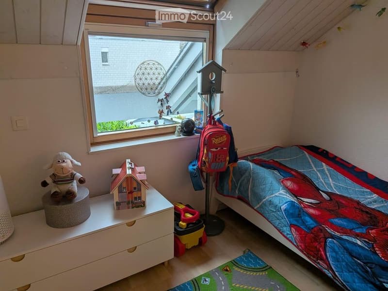 Cozy children's bedroom with a single bed, colorful Spider-Man bedding, a small toy house, a stuffed sheep toy, a backpack, and a toy car on the floor. The room features a large window with a view of the outside and a small shelf with additional toys.