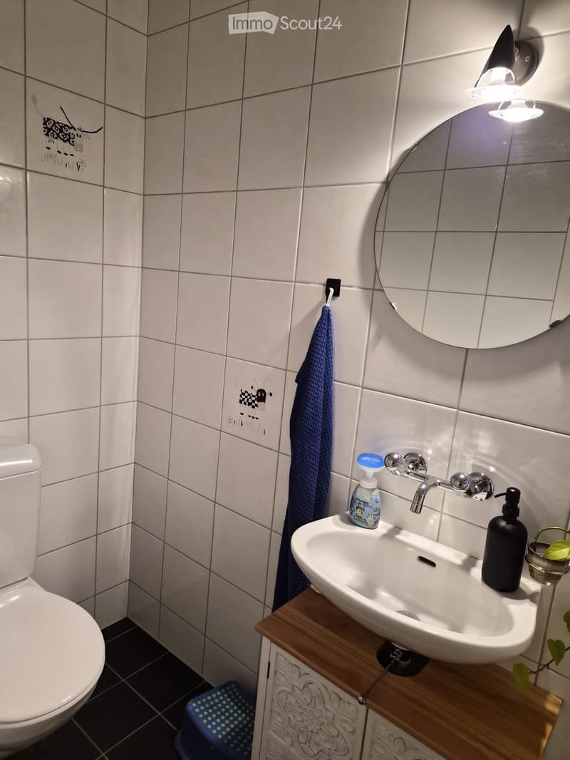 Bathroom with white tiled walls, a toilet, a sink with a wooden cabinet, a mirror, a towel, and a small stool.