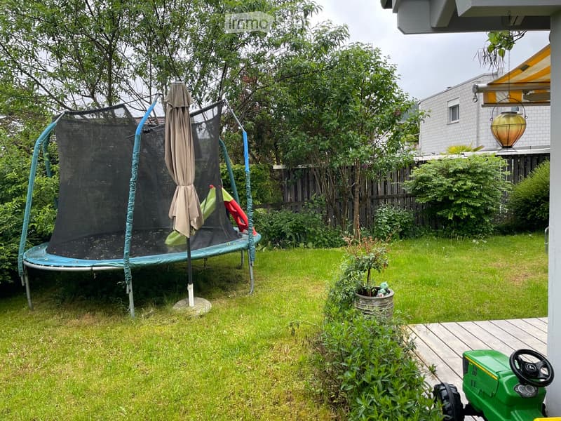 Garden with a trampoline, umbrella, small green tractor, potted plant, and surrounding greenery.
