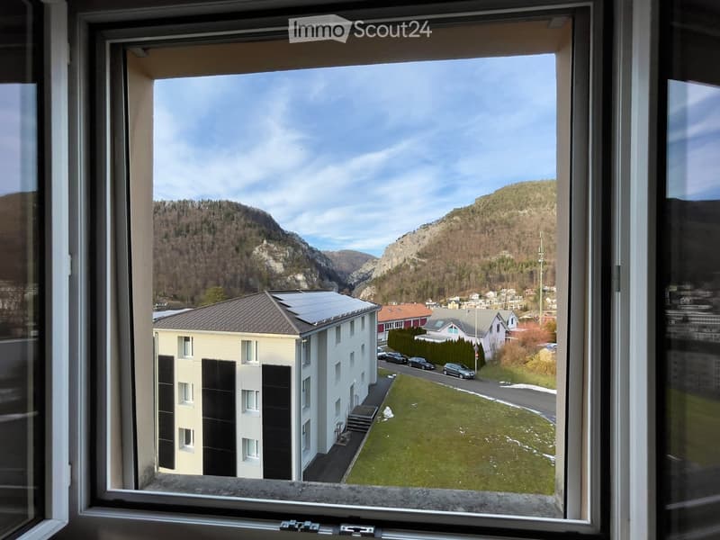 View from a window of an apartment overlooking a building with solar panels, grass, and a mountain range.
